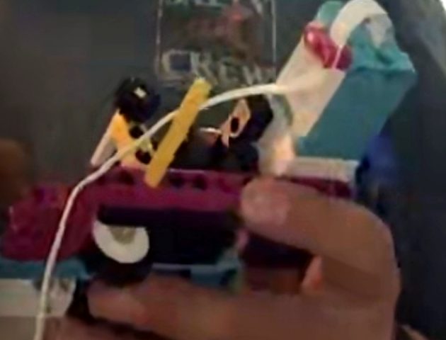 An early SPIKE assembly held in one hand: a motor, coloured Technic beams and a small wheel, with a cable attached.